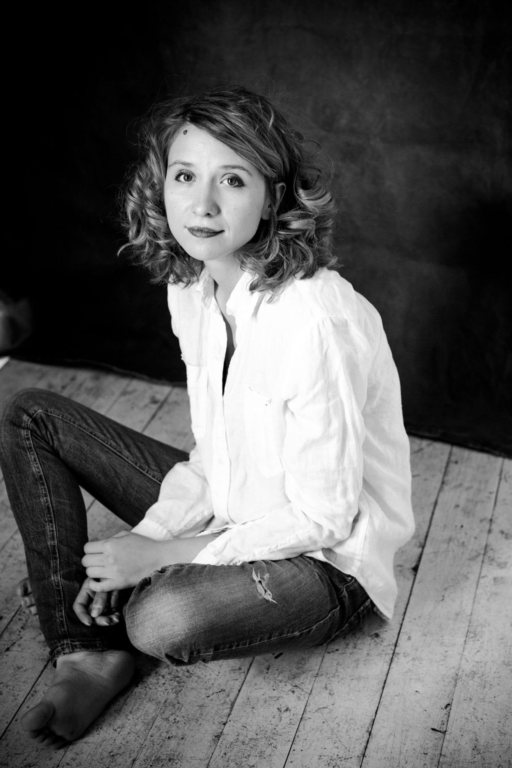 Alisa Kovalenko looking directly at the camera. She is sitting on a wooden floor and wearing denim trousers and a white shirt. Her shoulder-length hair is curly. Black and white portrait.