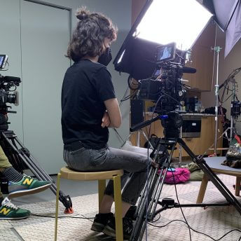 Production still of Hollywood Does Abortion. Director of Photography, Melissa Langer, is sitting behind camera gear looking at image of subject in viewfinder. Camera is on tripod and room is filled with lighting and camera gear.