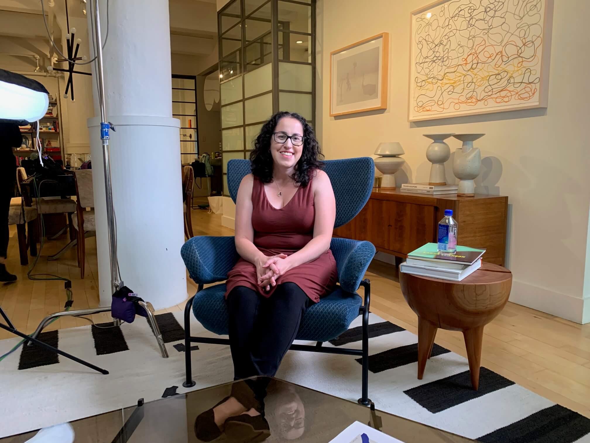 till from Hollywood Does Abortion. Steph Herold looking directly at the camera. She is wearing glasses and a sleeveless dress, sitting in a living room chair. WA light stand and light are additionally in the foreground.
