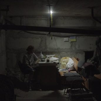 Production still of Intercepted. A woman is sitting at a table in profile to the camera. She is lit by a sole lightbulb and various bags of items can be seen to her right. While the frame is Some pipes are shown and concrete walls surround her. She is waiting in a bomb shelter underneath Kharkiv