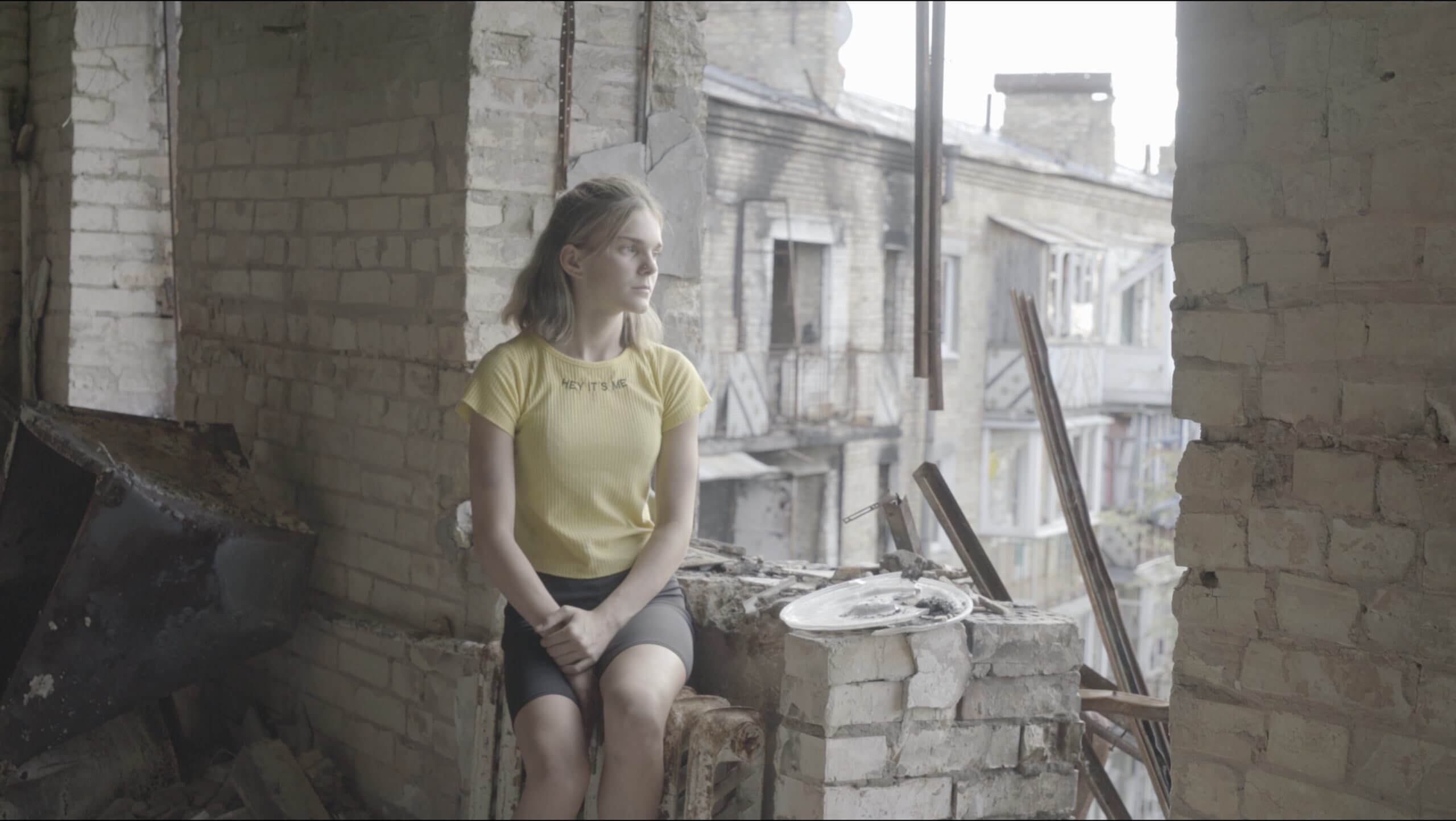 Production still of Intercepted. A young teenage girl in a yellow shirt is sitting in profile to the camera. She is on the edge of her former apartment's window. Surrounding her is rubble, the result of a recent bombing. In the background, the exterior of another bombed building can be seen.