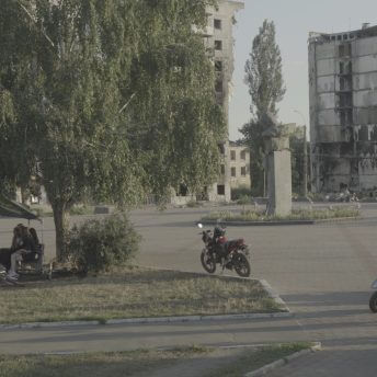 Production still of Intercepted. Two groups of people are gathering in a plaza. To the left, one group is sitting on benches underneath a large tree. A group of men to the right are standing near their motorbikes. Centered in the frame is a parked motorcycle. In the background are a damaged statue and a bombed out apartment building.