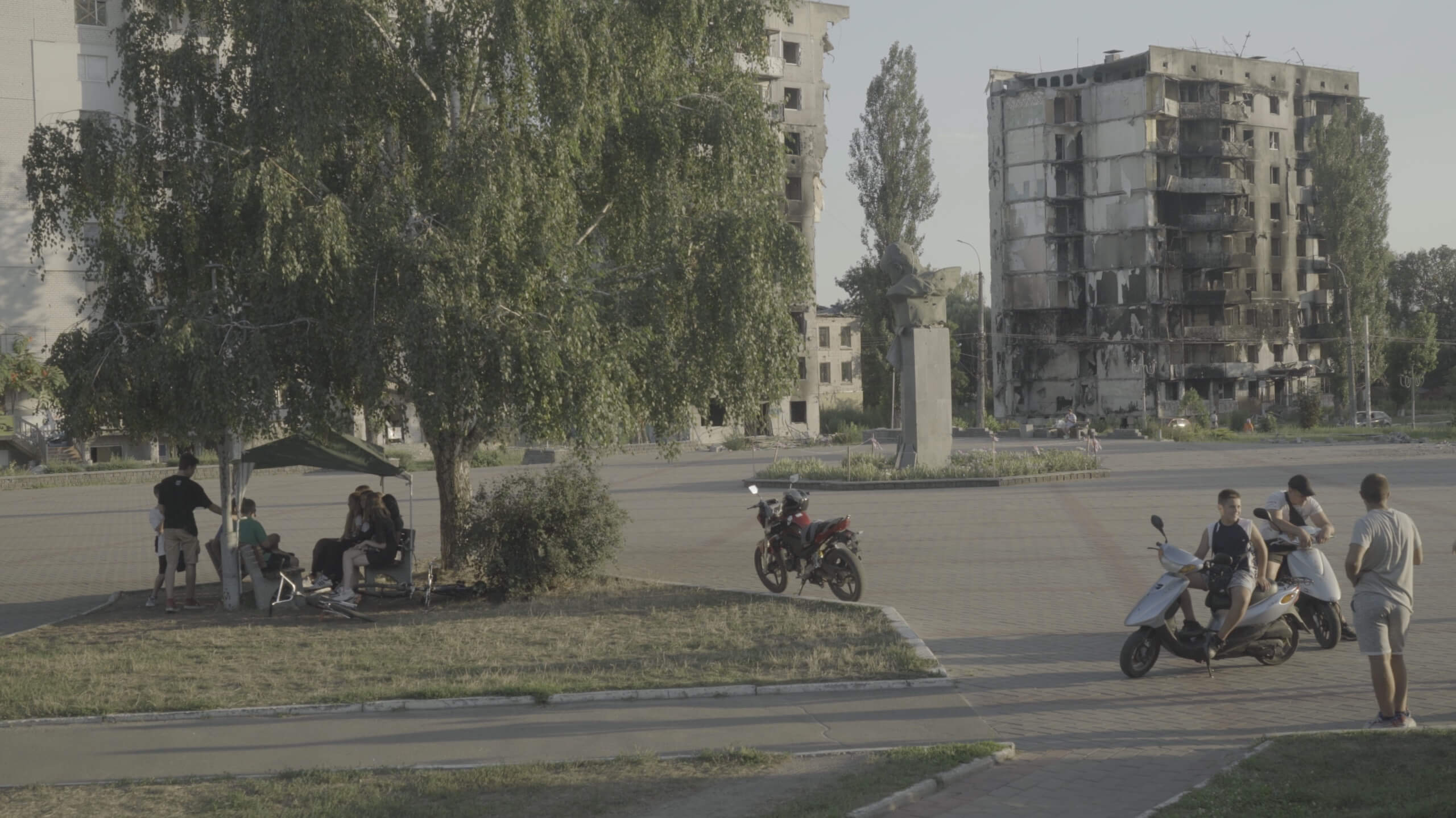 Production still of Intercepted. Two groups of people are gathering in a plaza. To the left, one group is sitting on benches underneath a large tree. A group of men to the right are standing near their motorbikes. Centered in the frame is a parked motorcycle. In the background are a damaged statue and a bombed out apartment building.