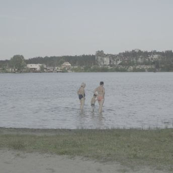 Still from Intercepted. A lake is shown from the shoreline. Various people in bathing suits are shown wading in the far left and right of the frame. Two women and their grandson are in the middle, with their backs to the camera. In the far background, across the lake, recently bombed buildings are visible.