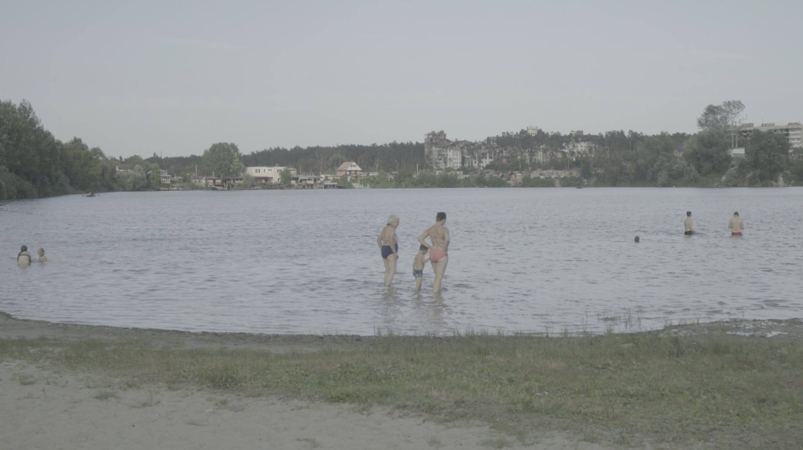 Still from Intercepted. A lake is shown from the shoreline. Various people in bathing suits are shown wading in the far left and right of the frame. Two women and their grandson are in the middle, with their backs to the camera. In the far background, across the lake, recently bombed buildings are visible.