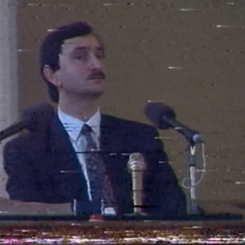 VHS archive image of a man in a dark suite and mustache standing behind microphones, his eyes skeptically overlooking behind the camera