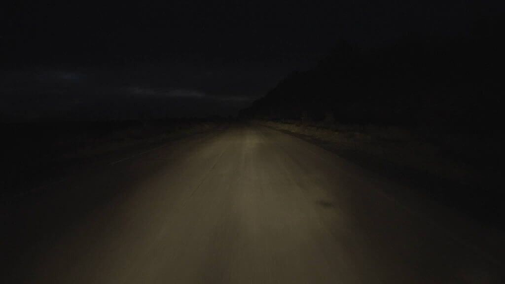 Still from Intercepted. A dark road is lit by a vehicle's headlights. A rural landscape can be seen; some vegetation on the left and right of the road; there are hills in the background and the sky's horizon is in the distance.