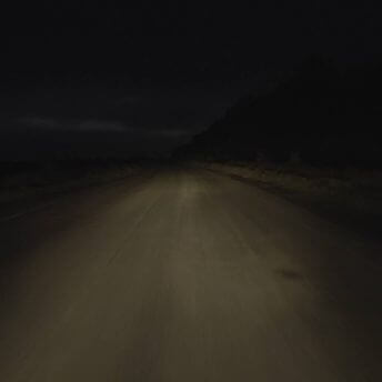 Still from Intercepted. A dark road is lit by a vehicle's headlights. A rural landscape can be seen; some vegetation on the left and right of the road; there are hills in the background and the sky's horizon is in the distance.