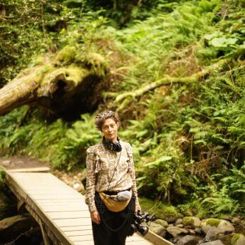 Filmmaker Salomé Jashi stands on the backdrop of greenery and a bridge, carrying the camera equipment