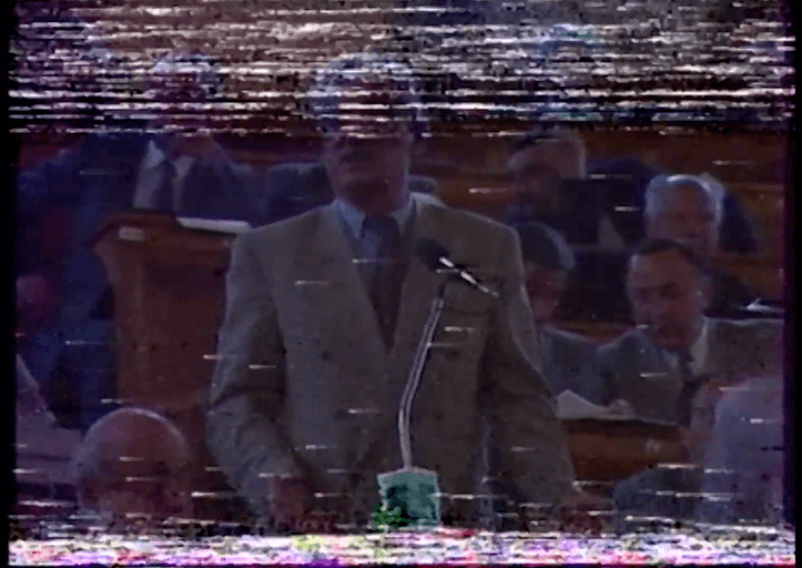 Damaged VHS archive image of a man in a white suite standing by a microphone in the Parliament. His head is covered with white noise. Other men in suites sit behind.