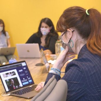 till from The Untitled 19th* News Film. A group of five women reporters sit around a table with masks on and laptops open to the 19th News digital website. The woman closest to the camera has her hand up to her mask.