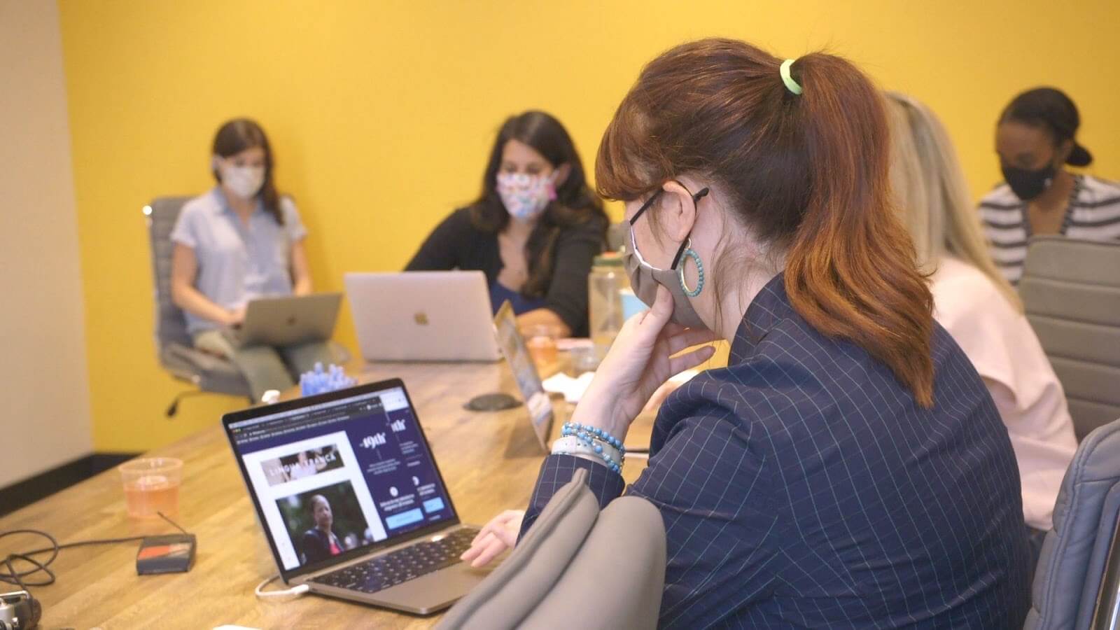 till from The Untitled 19th* News Film. A group of five women reporters sit around a table with masks on and laptops open to the 19th News digital website. The woman closest to the camera has her hand up to her mask.