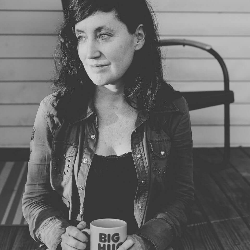 Heather Courtney looking away from the camera to her left. She has long, wavy hair and is wearing a jean shirt. She is holding a mug and sitting on a wooden porch. Black and white portrait.