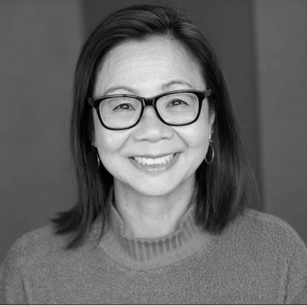 Diane Quon looking directly at camera and is smiling. She has mid-length hair and is wearing a turtleneck, gold hoop earrings and black rimmed glasses. Black and white portrait.