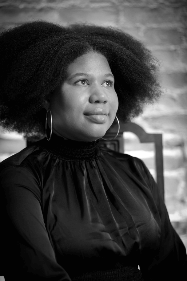 Princess Hairston looking off camera to her right. She has dark curly hair parted in the middle. She is wearing a black turtleneck blouse and large hoop earrings and is sitting in front of an out of focus brick wall. Black and white portrait.