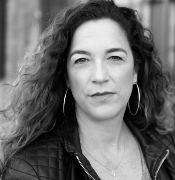 Producer Kristi Jacobson wears a leather jacket and hoop earrings. Her hair is light brown and curly. Black and white portrait.
