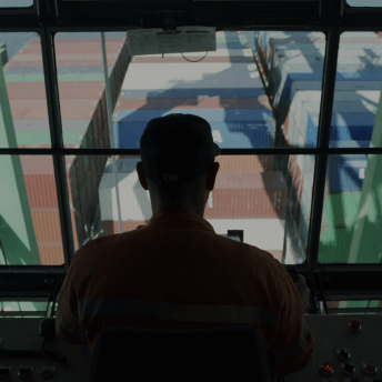 A profile of a man from behind as he sits in the control seat of a giant crane lifting shipping containers below.