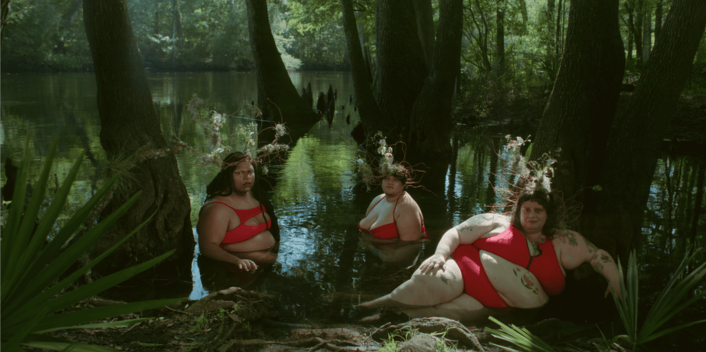 Still from How to Carry Water. Three people in swimsuits are posing for the camera. They are in a lagoon.