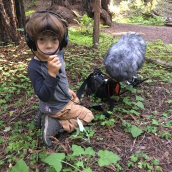 A young boy looks directly at the camera - his face is seen through a magnifying glass. He wears headphones and is sitting next to a sound recorder and boom microphone in the woods.