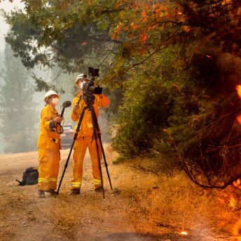 Two figures dressed in orange fire gear and masks look into a camera with flames in the foreground.