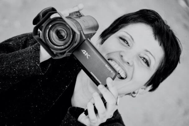 Maryam Ebrahimi smiles and points a camera directly at the screen. Black and white portrait.