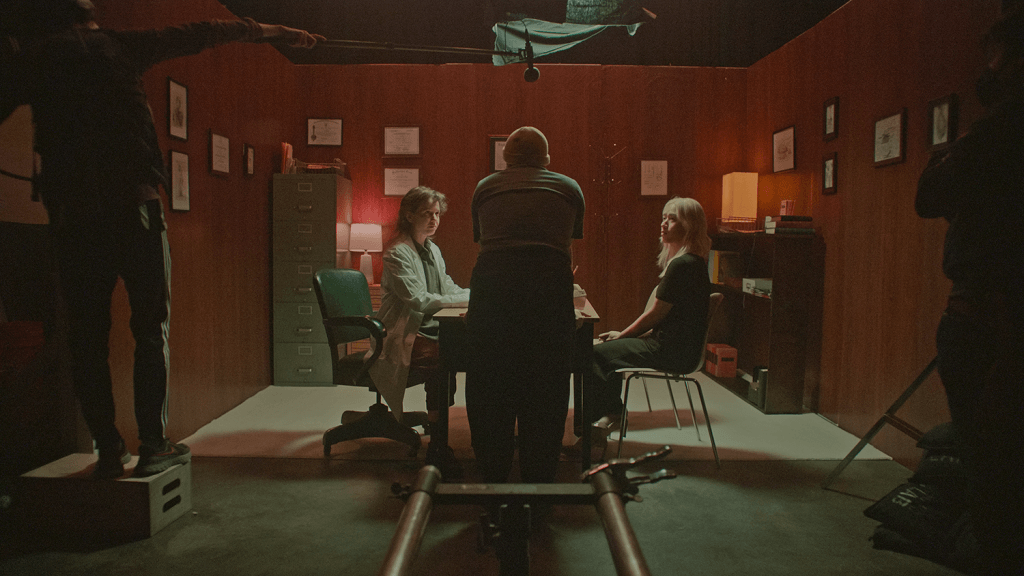 Still from The Script. In a set, a person with their back to the camera, talks to two other persons that are sitting in chairs in front of a table. To the right and left, there are camera crew people.
