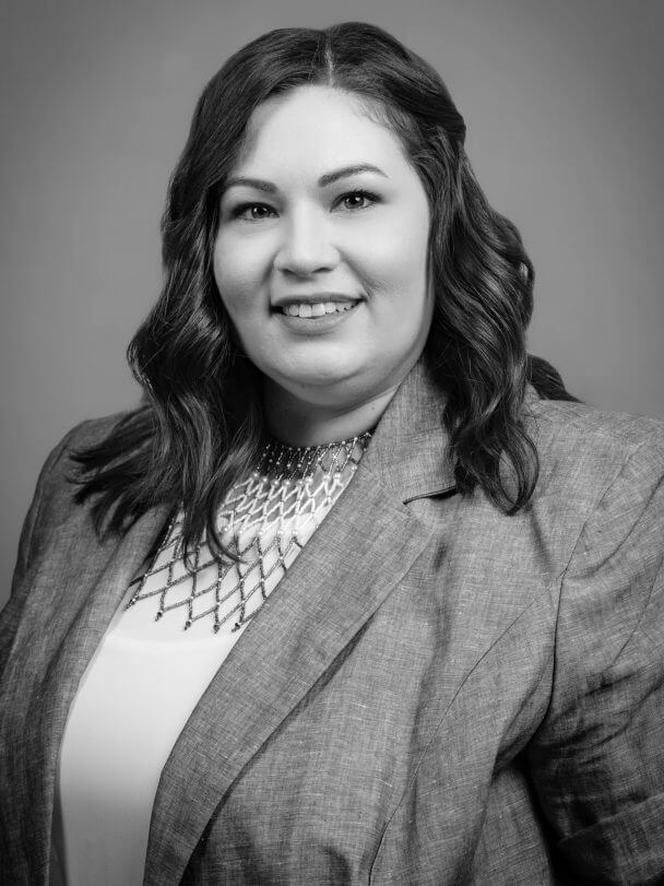 Rebecca Landsberry-Baker is an Indigenous woman with dark brown hair and light olive skin. She's wearing a beaded Muscogee collar and light blue jacket with a gray background. Black and white portrait.