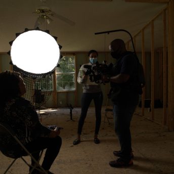 Two people stand with a camera in front of lights interviewing a seated Black woman.