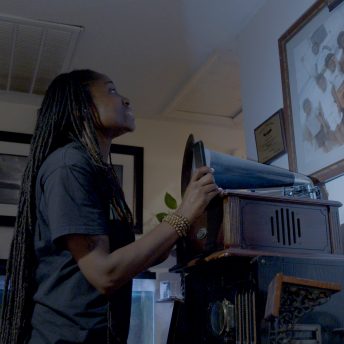 A Black woman with loc's looks up at a family portrait that is framed on the wall while standing in front of a old record player.