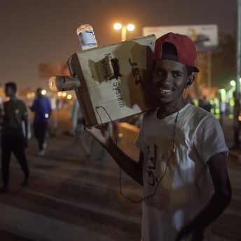 A Sudanese protestor smiles while carrying a cardboard camera on his shoulder.