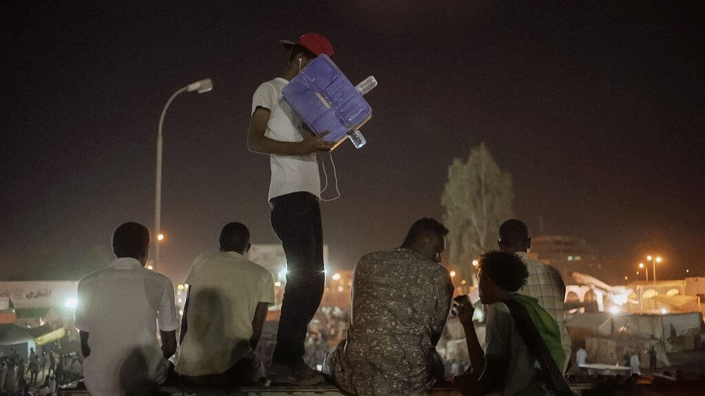 A young Sudanese protester points a cardboard camera towards an interview unfolding on a bridge in the middle of a large sit-in demonstration.