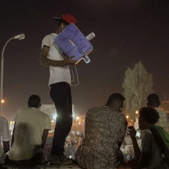 A young Sudanese protester points a cardboard camera towards an interview unfolding on a bridge in the middle of a large sit-in demonstration.