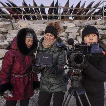 Director Lin Alluna, main character Aaju Peter and DoP Iris Ng shooting on location in Nuuk, Greenland, in front of a stand with many traditional Greenlandic kayaks.