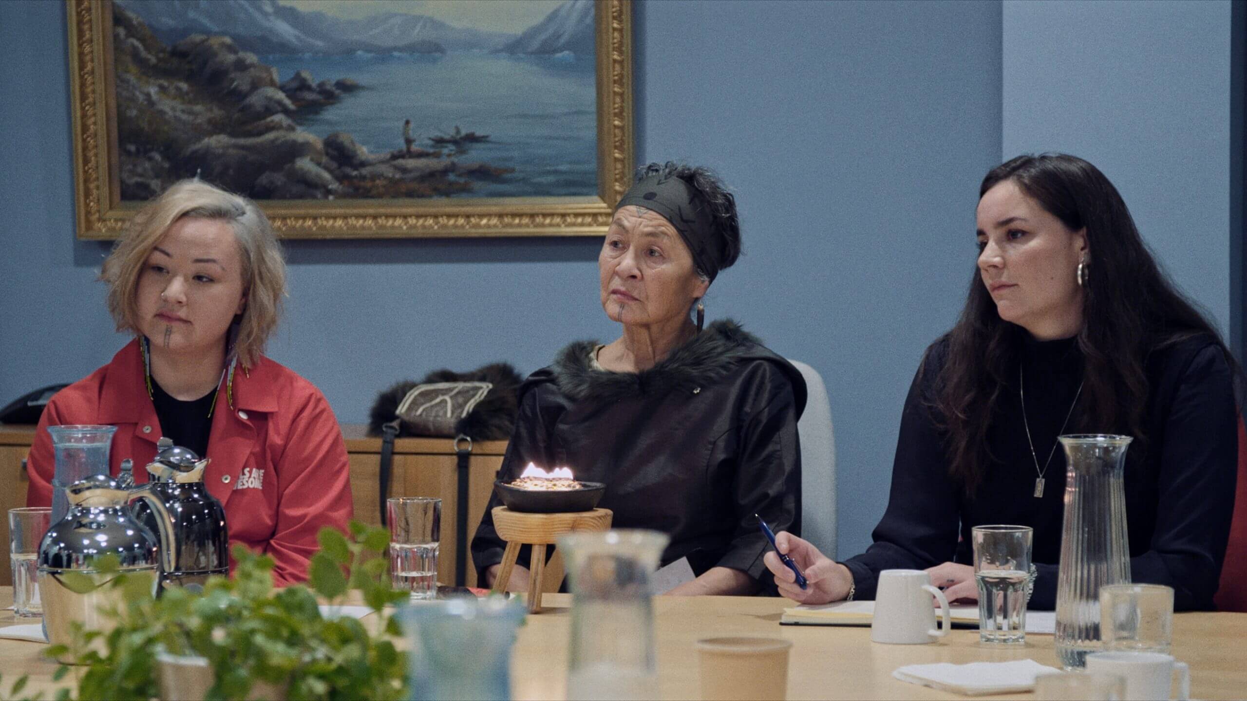 The main character of Twice Colonized, Aaju Peter, is in a meeting and is sitting next to two young fellow Inuit activist. There is a traditional Greenlandic oil lamp, a qulleq, in front of her, and all three women are looking serious but hopeful.