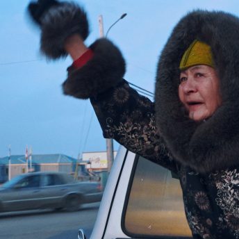 The main character of Twice Colonized, Aaju Peter, is in the back of a pick up truck in motion. She is wearing a fur coat, and she's waving enthusiastically.