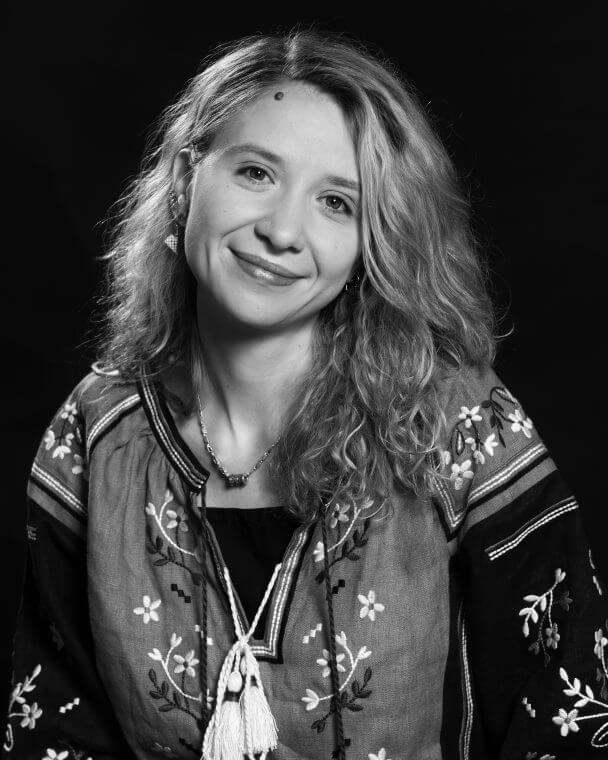 Alisa Kovalenko looks at the camera against a dark background and smiles. She is wearing a Ukrainian national embroidered shirt with flowers. The photo is in black and white.