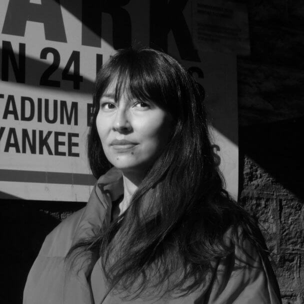Director Ursula Liang wears a red winter jacket and stands in dramatic shadows in front of a sign for a parking lot near Yankee Stadium. Black and white.
