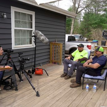 Director Tsanavi Spoonhunter conducting a sitdown interview with Menominee logger, Dustin Peters and his father, Alex Peters. They are seated outside of Dustin's home on a summer day.