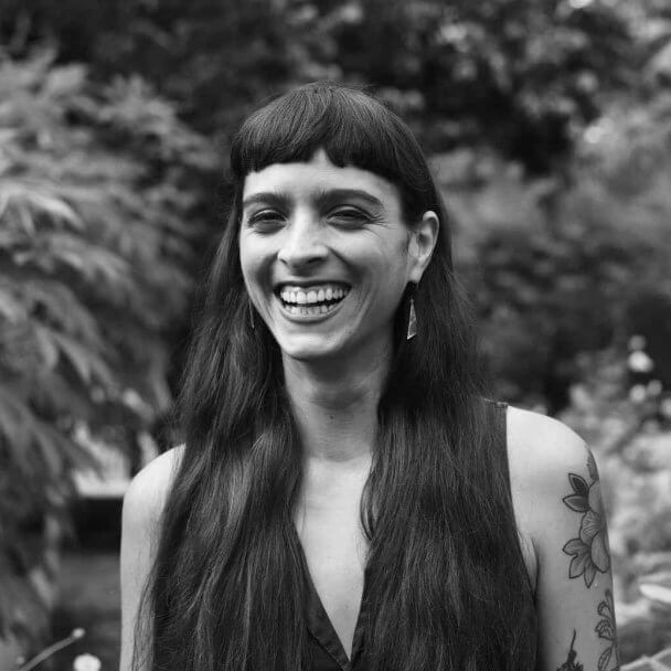 One of the producers for Matininó with long brown hair, bangs, orange eyeshadow and tattoos smiles for a photograph behind a backdrop trees.