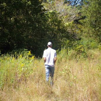 a Black man with a light hat walks through a vacant lot with overgrown weeds