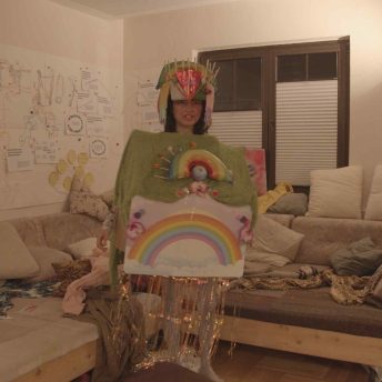 In a messy lounge room, Viv stands alone, looking confused, in a rainbow house costume made out of cardboard.