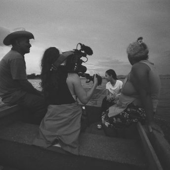 A woman has a camera rigged on her shoulder and films a small group of four as they all sail a lake on a small motor boat.