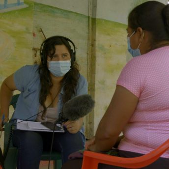 A woman wearing jeans, a blue top and a face mask, holds a microphone, sits, takes notes and interviews another woman, who has her back turned towards the camera and is wearing a pink blouse.
