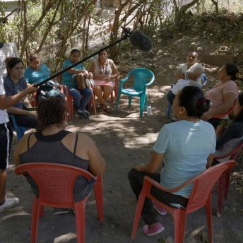 A group of women sit outdoors in a circle and have a discussion, while a person stands and holds a boom as they record the conversations.
