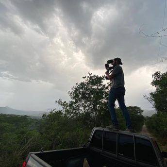 A cameraman stands on top of a pick-up truck and films the mountainous landscape
