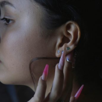 A young woman tucks a hair strand behind her ear with her acrylic nails, painted with bright pink and rose pink nail polish and decorated with glitter.