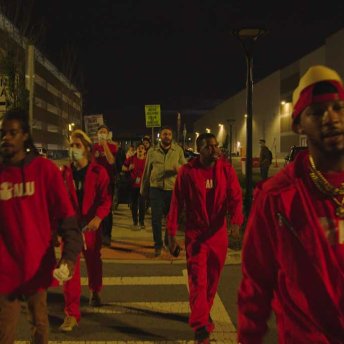 Still from Union. A group of people wearing red walk in front of the camera. It is night time. Some of them wear facemasks.