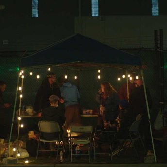 Still from Union. A group of people stand under a tent covered with a string light. It is night time. Some of them are sitting.