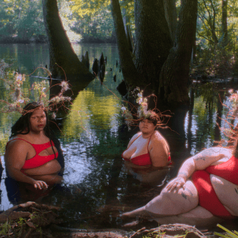 Three fat, gender non-conforming people of different races pose in a lush swamp wearing striking red bathing suits and crowns made of plants.