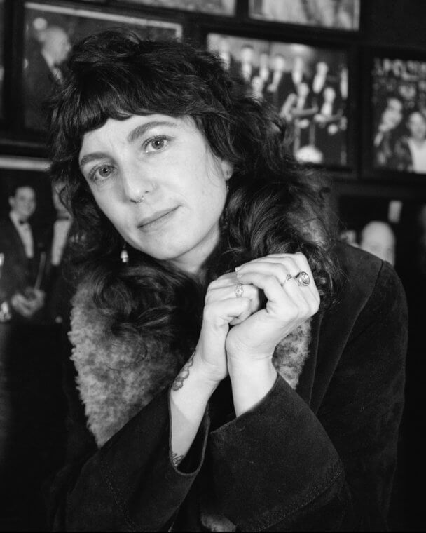 A white woman with wavy brown hair in a fur trimmed coat and looks at the camera with their hands clasped.